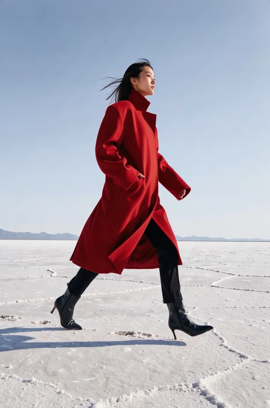 Editorial fashion photograph of a model in an oversized sculptural red wool coat walking across a white salt flat, wide shot, high fashion magazine